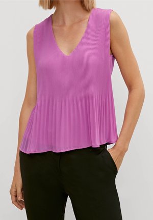 Haut sans manches violet plissé à col en V porté avec un pantalon noir, main droite du mannequin dans la poche, fond neutre.