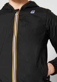 Veste noire à capuche avec une fermeture éclair rayée multicolore (jaune, blanc, marine) et un petit logo rouge, blanc et bleu sur la poitrine.