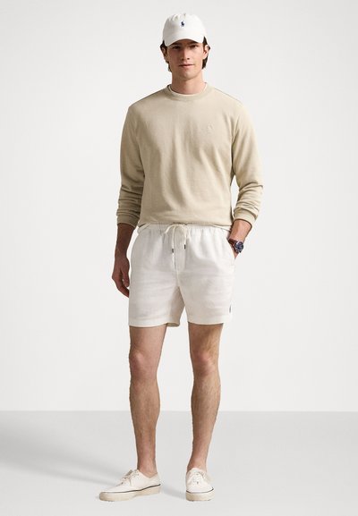 Beige hosszú ujjú ing, fehér zsinórderekes rövidnadrág és fehér sneakers. Kiegészítők között szerepel egy fehér sapka és egy kék számlapú óra.