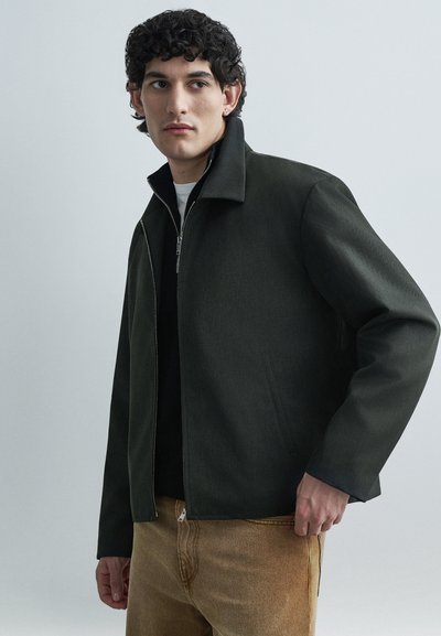 Veste vert foncé en tissu texturé, dotée d'une fermeture éclair, d'un col avec revers et de poches latérales, portée sur un haut noir.