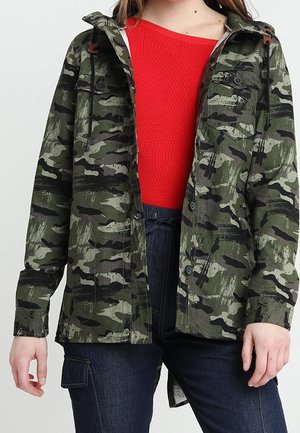 Personne portant une veste camouflage verte et noire sur un pull rouge vif et un jean denim bleu foncé avec une poche cargo.
