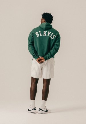 Hombre de pie con las manos entrelazadas detrás, vistiendo una sudadera con capucha verde con el texto "BLKVIS", pantalones cortos blancos con rayas finas, zapatillas blancas Nike y gafas.