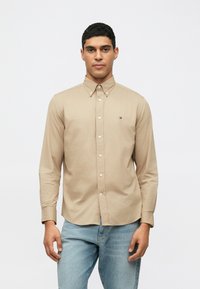 Camicia a maniche lunghe color beige con colletto, caratterizzata da un piccolo logo sul petto, abbinata a jeans in denim azzurri.