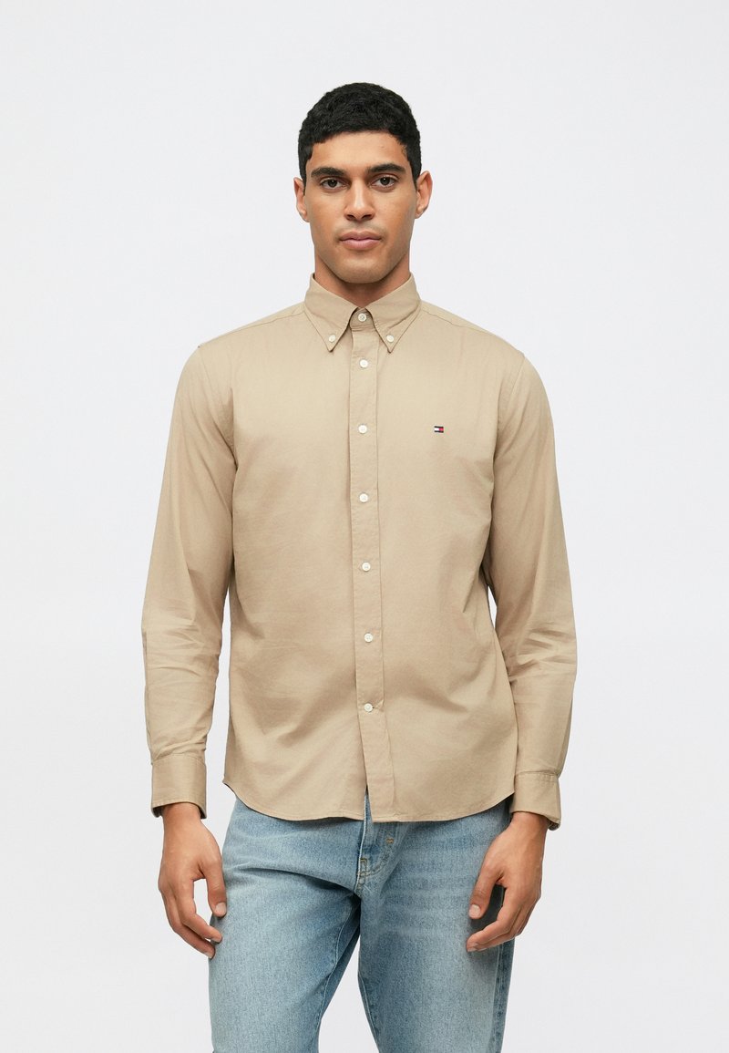 Camicia a maniche lunghe color beige con colletto, caratterizzata da un piccolo logo sul petto, abbinata a jeans in denim azzurri.