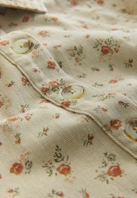 Tkanina w beżowym kolorze z kwiatowym wzorem składającym się z małych czerwonych i zielonych kwiatów, wyposażona w dwa kremowe guziki i teksturowane wykończenie.