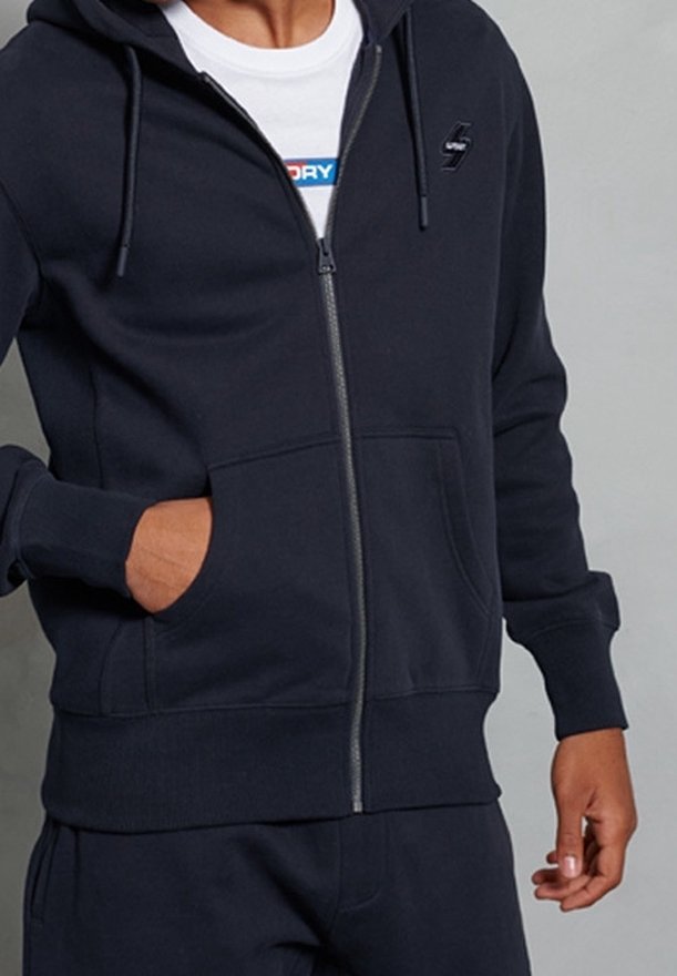 Personne portant un sweat à capuche noir zippé avec les mains visibles, montrant un t-shirt blanc avec du texte bleu et rouge en dessous.