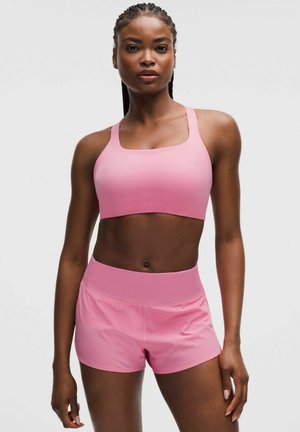 Atletisk kvinde med fletninger iført en lyserød sports-bh og matchende lyserøde træningsshorts, stående mod en ensfarvet grå baggrund.
