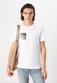 T-shirt en coton blanc avec un graphique rectangulaire en gris et noir. Le modèle tient un sac à épaule en suède beige. Coupe décontractée et jeans bleu clair.