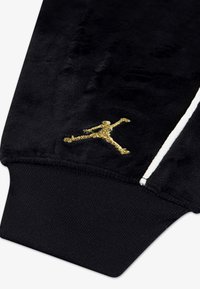 Čierny velúrový rukáv s rebrovaným manžetovým zakončením, s vyšitým zlatým logom Jumpman a bielym akcentovým prúžkom pozdĺž švu.