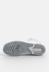 Białe buty sportowe z teksturowaną gumową podeszwą z geometrycznym wzorem bieżnika oraz logo "new balance" na boku.