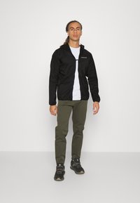 Black lightweight jacket with a zipper, hood, and logo, paired with olive green pants and black shoes, displayed on a neutral background.