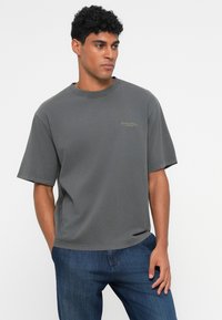 T-shirt in cotone grigio con maniche corte, colletto rotondo e un discreto logo sul petto. Indossato con jeans blu. Design semplice e vestibilità rilassata.