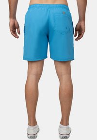 Pantaloni da bagno elastici blu con due tasche laterali e una tasca posteriore. Texture liscia con un patch del logo sul retro. Indossati con sneakers bianche.