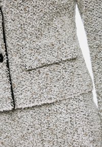 Strukturiertes Stoffanzug in Off-White und Beigetönen. Verfügt über einen schmalen Revers und eine Seitentasche mit schwarzem Besatz an den Kanten.