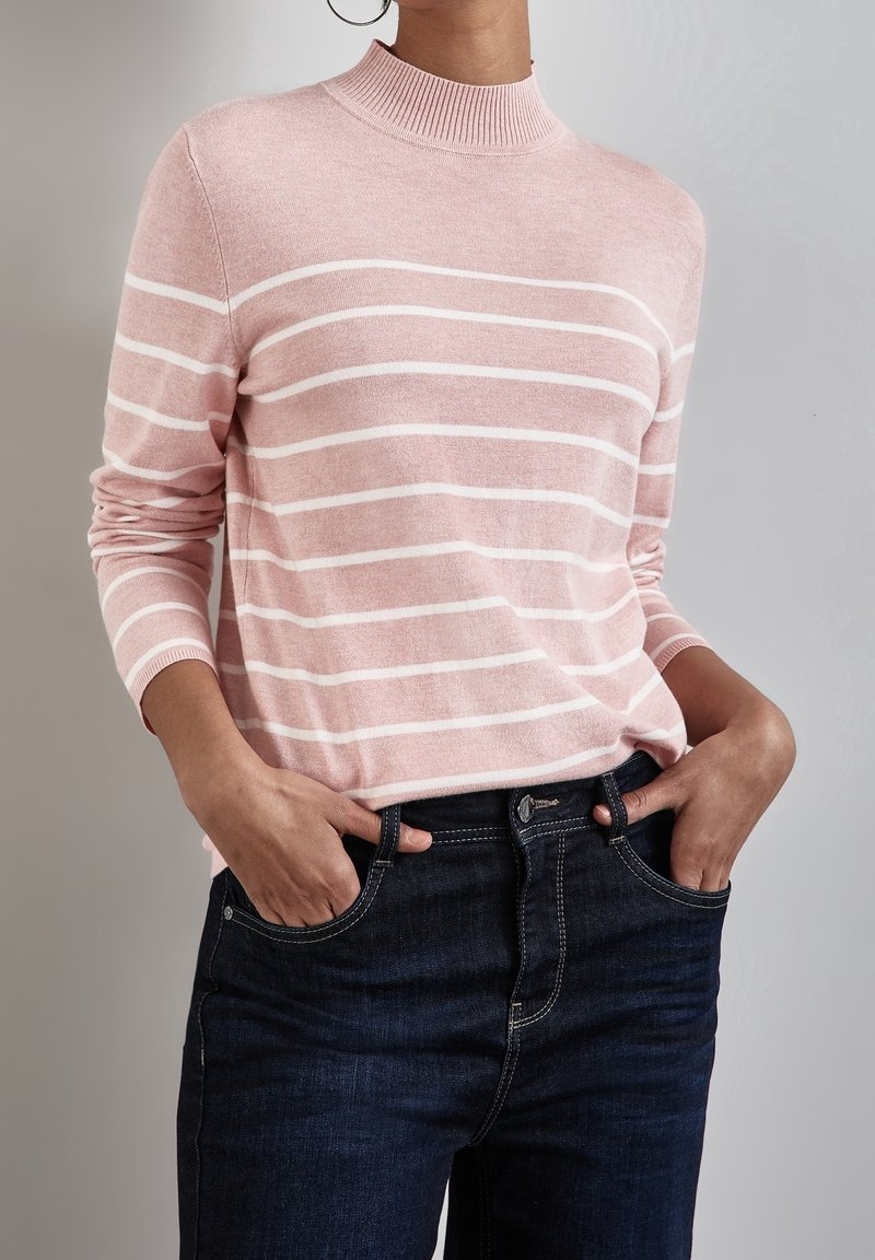 Maglione in maglia rosa chiaro con righe orizzontali bianche, collo alto, maniche lunghe, abbinato a jeans blu scuro. Texture morbida, vestibilità comoda.