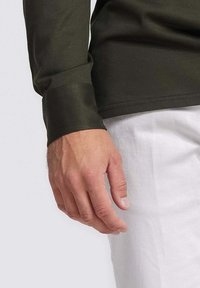 Close-up van een hand die een donkergroene lange mouw shirt draagt, met een gladde textuur en manchet, naast lichtgekleurde broeken.