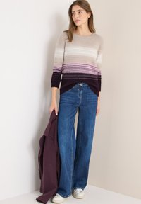 Pullover lavorato a maglia con larghe righe beige, rosa e viola, abbinato a jeans blu larghi e sneakers bianche. È indossato un cappotto morbido e texture.