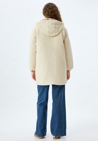 Krämfärgad, plysch fleecejacka med huva, långa ärmar och en avslappnad passform. Buren över blå utsvängda jeans och beige skor, sedd bakifrån.