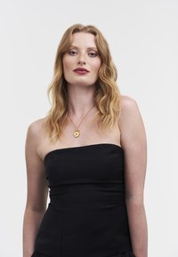 Musta olkaimeton toppi, jossa on istuva malli, sileä pinta ja hillittyjä yksityiskohtia. Mallilla on kaulassa kultainen riipusketju.