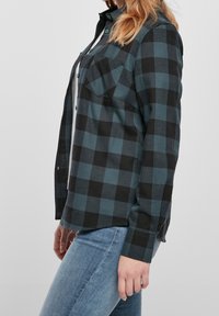Camicia di flanella a bottoni in verde scuro e modello a quadri neri, con due tasche sul petto e orlo dritto, realizzata in un tessuto testurizzato.