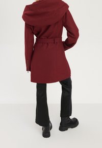 Abrigo burdeos con capucha y cinturón en la cintura, mangas largas y bolsillos laterales con cremallera. Combinado con pantalones de pierna ancha negros y botas negras gruesas.