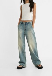 Top corto blanco combinado con pantalones vaqueros de pierna ancha de color azul claro. Los vaqueros tienen un lavado desgastado y bolsillos laterales. Sandalias planas negras completan el conjunto.