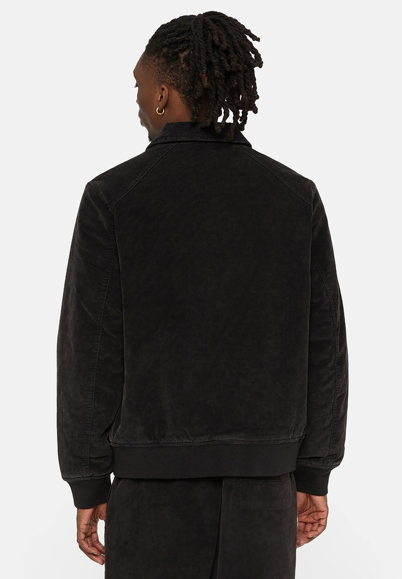 Veste aviateur noire avec une finition texturée, des poignets côtelés et un col. Présente une coupe décontractée, des coutures visibles et un design arrivant à la taille.