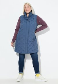 Blau gesteppte ärmellose Weste mit hohem Kragen, Frontreißverschluss und seitlichen Taschen, über einem weinroten Langarmoberteil und blauen Jeans. Weiße Sneaker.