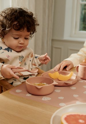 Tout-petit aux cheveux bouclés assis à une table, tenant une cuillère au-dessus d'un bol de nourriture, avec des tranches de melon et une tasse à proximité sur une nappe.