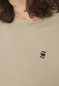 Primo piano di una persona che indossa una maglietta beige con un piccolo logo nero G-Star RAW sul petto, mostrando spalla e collo con capelli castani.