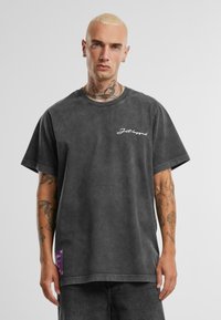 Czarny, oversize T-shirt wykonany z miękkiego, teksturowanego materiału, z białym, kursywno zapisanym logo oraz fioletową metką na dole. Krótkie rękawy.