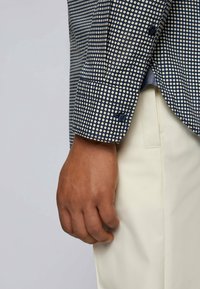 Granatowa koszula w białe kropki, z mankietem na guziki i teksturowaną tkaniną. Noszona z jasnymi spodniami. Ręka widoczna na krawędzi.