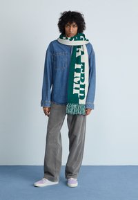 Camisa de ganga, calças de cortiça cinzentas, sapatos em roxo pastel e um lenço verde/creme com franjas e texto em negrito "VIAGEM". Fundo plano.