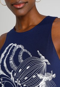 Granatowa, bezrękawnikowa bluzka z białym wzorem kwiatowym oraz zakrzywionymi, teksturowanymi liniami. Materiał ma gładkie wykończenie i dopasowany krój.