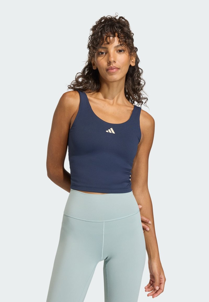 Vrouw draagt een donkerblauw mouwloos crop top met een wit logo en lichtblauwe leggings met hoge taille, staat met één hand achter haar rug.