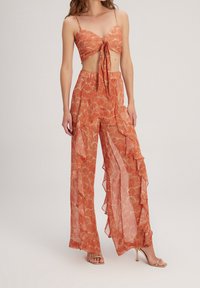 Conjunto de dos piezas que incluye un top corto de estampado naranja con un frente anudado y pantalones amplios de pierna ancha, transparentes y con volantes que fluyen de manera suelta.