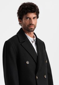Abrigo negro de lana con corte cruzado, presenta un solapa de punta, dos botones en tono dorado y un bolsillo lateral, combinado con una camisa de rayas claras.