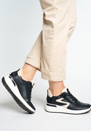 Person trägt beige Cordhosen und schwarz-weiße Sneaker, mit einem leicht angehobenen Fuß vor einem schlichten hellen Hintergrund.