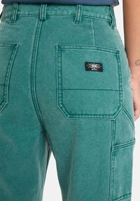 Pantalons cargo teal en tissu léger, avec plusieurs poches utilitaires et une étiquette avec un emblème noir sur la poche arrière.