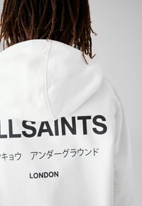 Fehér kapucnis pulóver, amelyen fekete szöveg olvasható: "ALLSAINTS" és "LONDON" a hátsó részen. Puha anyagból készült, laza szabással.