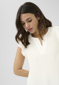 Blouse blanche à manches courtes avec un tissu texturé, présentant un décolleté en V et des accents de volants sur les manches. Design simple et élégant.