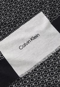 Sciarpa nera con motivo a logo ripetuto in bianco, dotata di un'etichetta bianca a contrasto con la scritta "Calvin Klein" in nero, bordi frangiati.