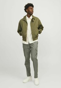 Chaqueta bomber verde oliva, camisa blanca de botones, pantalones cargo gris claro y zapatillas blancas. Tejido suave, corte relajado, diseño minimalista.