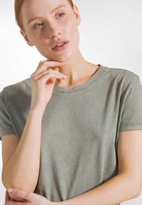 Ljusgrön bomulls-t-shirt med rund hals, korta ärmar och mjuk textur. Modellen poserar eftertänksamt, med en hand nära hakan.