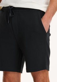 Schwarze Baumwollshorts mit einem Kordelzugbund, seitlichen Taschen und genähten Säumen, die eine glatte Textur und eine lockere Passform bieten.