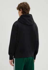 Felpa nera con cappuccio, realizzata in tessuto morbido. Presenta polsini e orlo a coste, con una vestibilità comoda, mostrata da dietro.