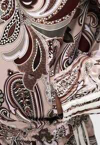 Gros plan d’un bras plié portant un vêtement à manches longues avec un motif cachemire marron, beige et blanc et un poignet élastiqué.