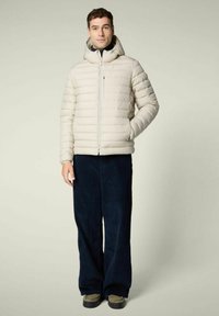 Uomo che indossa un giubbotto imbottito con cappuccio beige chiaro, pantaloni larghi scuri e sneakers, in piedi su uno sfondo chiaro e semplice.