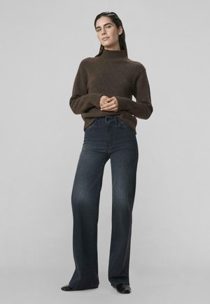 Frau mit dunklem Haar trägt einen braunen Rollkragenpullover, hoch geschnittene dunkelblaue weite Jeans und schwarze Schuhe, steht vor einem einfarbigen Hintergrund.