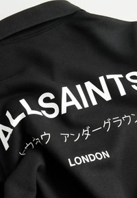 Čierna mikina s bielym nápisom "ALLSAINTS" a ďalšími japonskými znakmi, s klasickým golierom a jemnou textúrou.